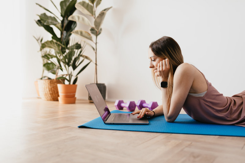 Frau liegt auf einer Sportmatte und schaut Webinar am Laptop